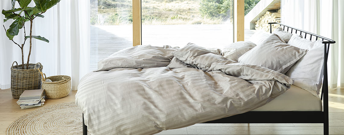 Spavaća soba s crnim metalnim krevetom, poplunima i jastucima, prekrivena prugastom posteljinom u svijetlosivoj i bijeloj boji