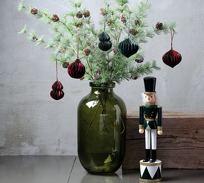 Zelena staklena vaza napunjena s umjetnim grančicama sa šišarkama, božićni ukrasi u zelenoj i crvenoj boji. Pokraj stoji božićna figurica